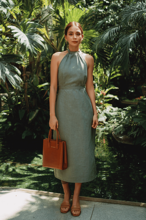 woman in a sophisticated halter-neck midi dress in muted sage linen, with a subtle A-line silhouette and minimal seam detailing, paired with tan leather block-heel sandals