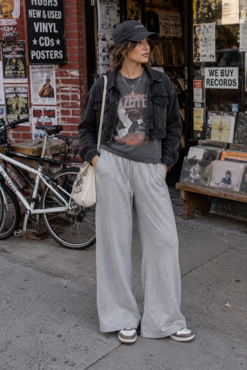 A woman wears a black baseball cap with a cropped black denim jacket, a vintage graphic T-shirt, wide-leg gray sweatpants, and gray-and-white sneakers