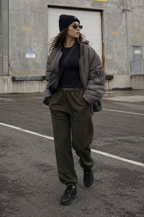 A woman wears a black beanie with an oversized gray bomber jacket, a fitted black top, olive green sweatpants, and chunky black sneakers