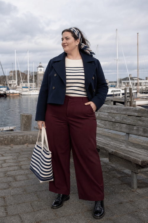 Plus-size woman wearing a navy peacoat, striped sweater, burgundy trousers, loafers, and a striped tote.