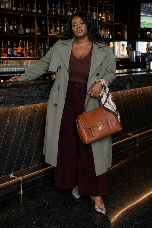 Plus-size woman wearing an olive trench coat with burgundy wide-leg pants and a brown leather handbag.