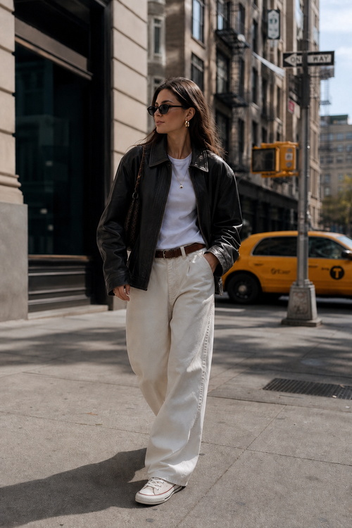 Woman in a black leather jacket, white trousers, and Converse sneakers on a city street.