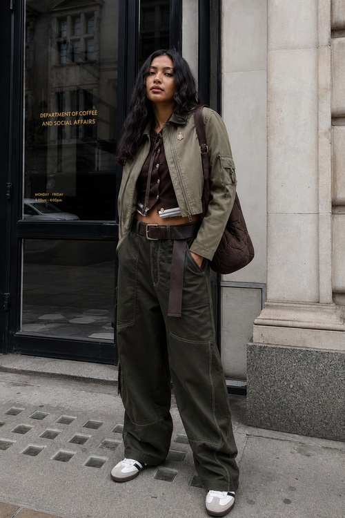 Woman in an olive cropped jacket, dark cargo pants, brown cardigan, and a quilted brown shoulder bag standing on a city street.