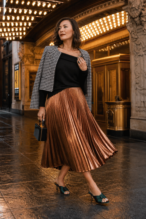 Woman wearing a black-and-white houndstooth cropped jacket over a black knit top with a bronze metallic pleated midi skirt, teal heeled sandals, and a structured top-handle bag