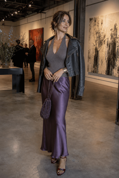 Woman wearing a black leather moto jacket over a taupe halter bodysuit with a lavender satin maxi skirt, embellished heeled sandals, and a beaded evening pouch inside a contemporary art gallery
