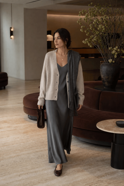 Woman wearing a gray satin slip dress layered with a cream cardigan, dark brown ballet flats, a charcoal scarf, and a brown leather handbag.