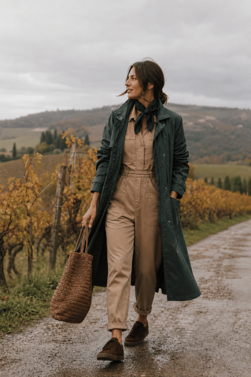 Woman wearing a green raincoat, tan utility jumpsuit, neck scarf, sneakers, and a woven tote.