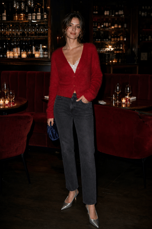 Woman wearing a red mohair cardigan over a lace-trim camisole with charcoal straight-leg jeans, silver pointed-toe pumps, and a navy velvet clutch inside a cozy cocktail bar