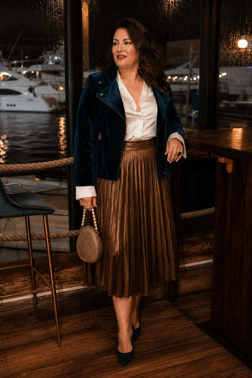 Woman wearing a teal velvet moto jacket with a bronze pleated midi skirt at a waterfront restaurant.