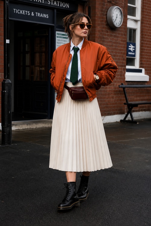 a woman wears a fall outfit with a rust bomber jacket, white pleated skirt, green tie, black combat boots, and a burgundy belt bag