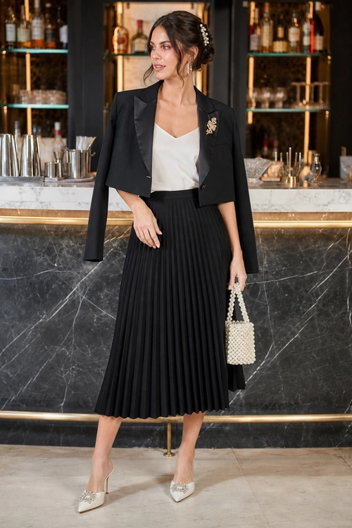 Elegant outfit featuring a cropped black blazer, white camisole, pleated black midi skirt, embellished ivory shoes, and a beaded handbag.