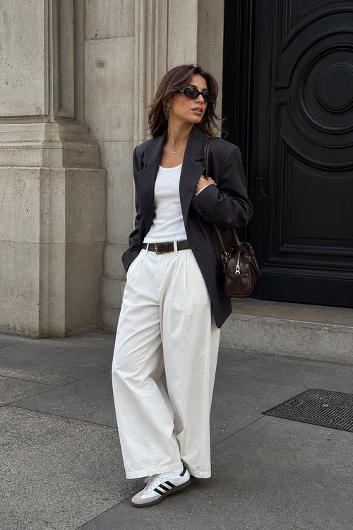 Black Blazer and White Wide-Leg Pants A woman wears a black blazer over a white top with white wide-leg pants, striped sneakers, sunglasses, and a brown shoulder bag.
