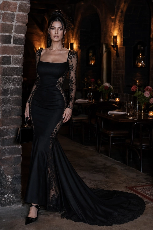 A woman wears a fitted black mermaid gown with sheer lace sleeves, lace panels, a long train, black heels, and a small black handbag.