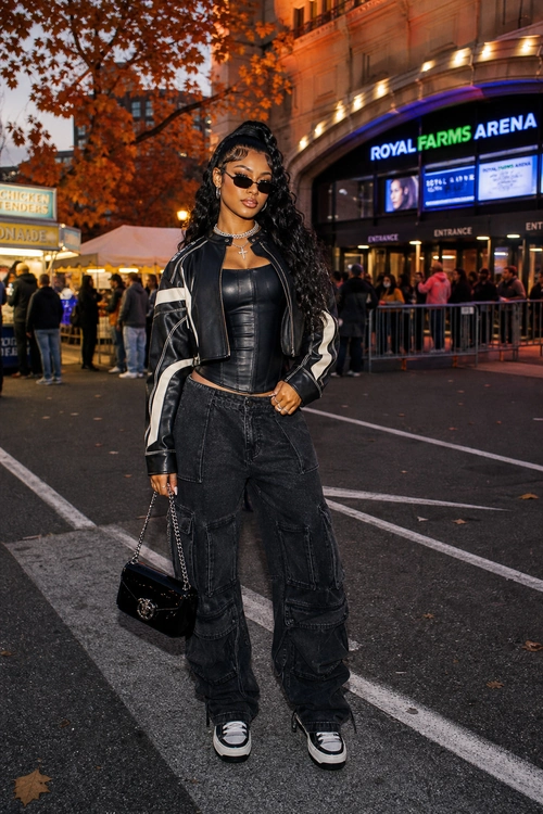 A woman wears a cropped black jacket with white stripes, a fitted black top, loose cargo jeans, black and white sneakers, sunglasses, and a chain-strap bag.