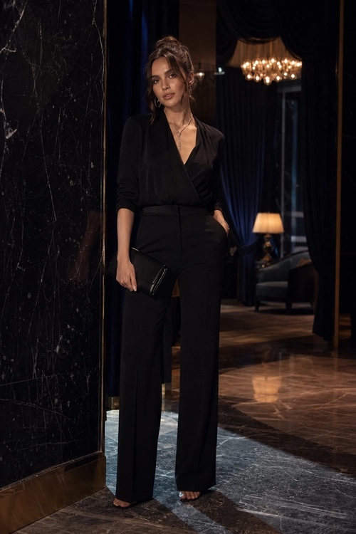 A woman wears a black wrap blouse with wide-leg trousers, a black clutch, a necklace, and open-toe sandals.