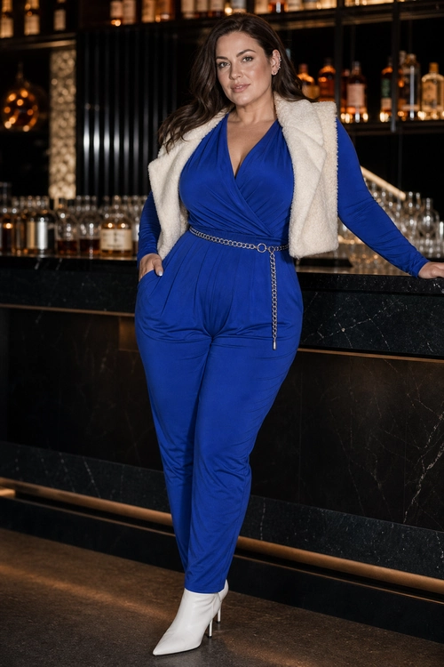 A royal blue wrap-style jumpsuit is styled with an ivory faux fur vest, a silver chain belt, and white pointed ankle boots.