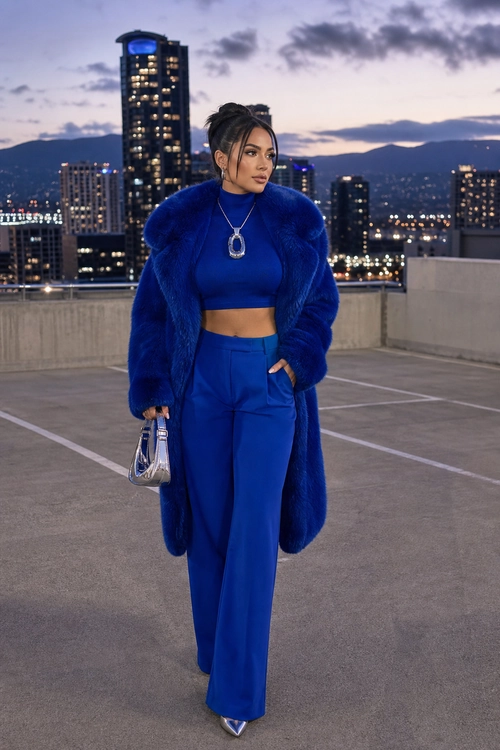 A woman wears a vivid blue cropped top, wide-leg trousers, and plush long coat with silver accessories on a rooftop at dusk.