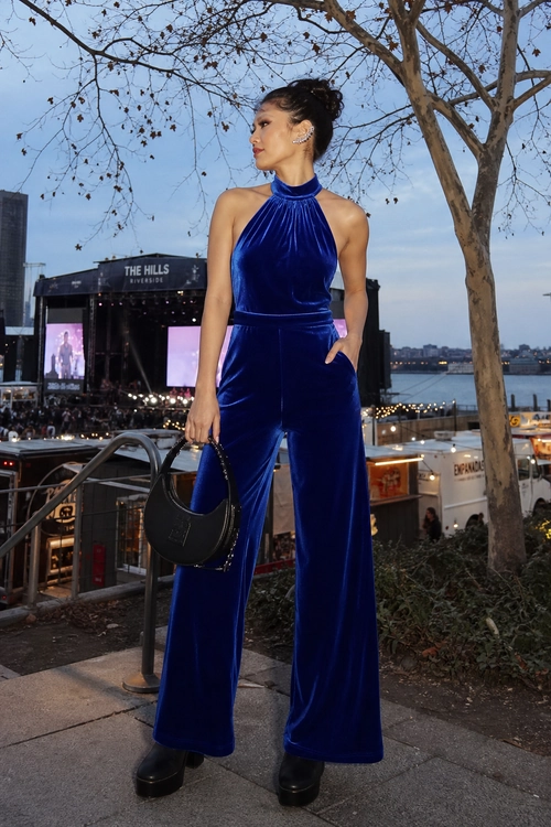 A woman wears a royal blue velvet halter jumpsuit with wide legs, black platform boots, and a black crescent handbag.