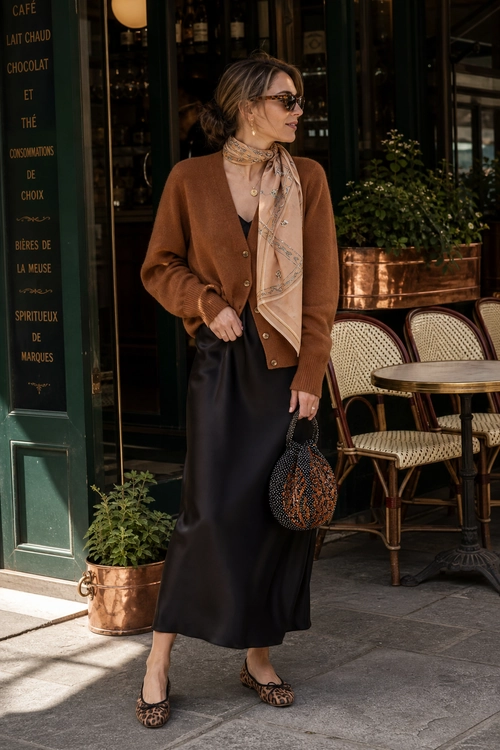 Brown Cardigan and Satin Skirt A woman wears a brown button-front cardigan with a black satin skirt, beige neck scarf, sunglasses, leopard flats, and a small patterned handbag.