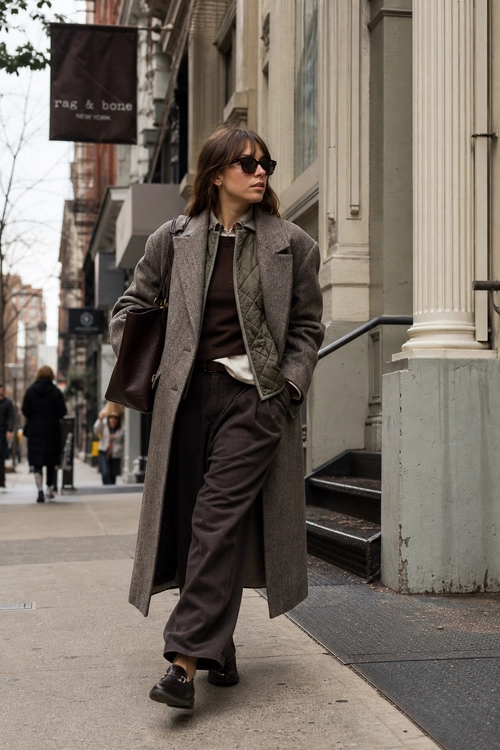 A person wearing a long gray coat, quilted vest, dark brown sweater, wide-leg trousers, sunglasses, and black loafers on a city sidewalk.