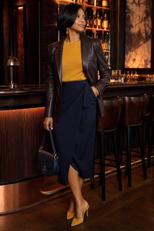 Leather Blazer and Wrap Skirt A woman wears a dark brown leather blazer with a mustard knit top, navy wrap midi skirt, yellow heels, gold jewelry, and a navy handbag.