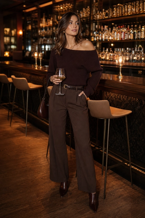 A woman wears a dark brown off-the-shoulder sweater with brown wide-leg trousers, a belt, pointed ankle boots, and a shoulder bag in a bar.