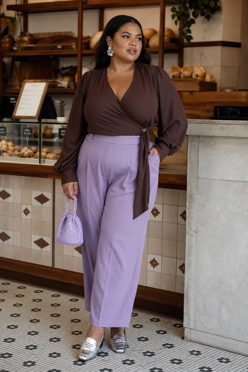 A woman wears a brown wrap blouse with lavender wide-leg trousers, a small lavender bag, silver loafers, and statement earrings.