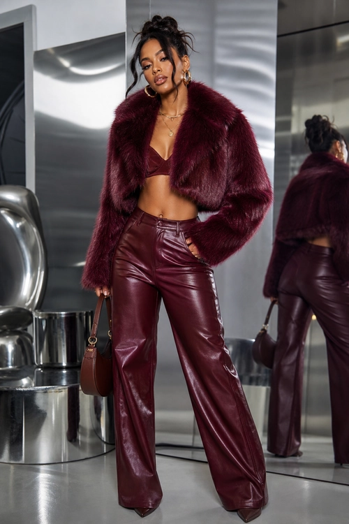 A woman wears a burgundy cropped fur-textured jacket, matching bralette, glossy wide-leg pants, pointed heels, and a small brown handbag.