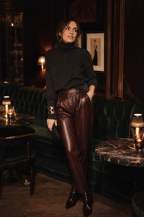 A woman wears a dark turtleneck sweater with burgundy leather trousers, pointed ankle boots, a metallic cuff bracelet, and a small black clutch.