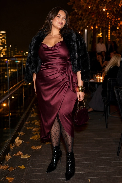 A woman wears a burgundy satin wrap dress with a black faux fur jacket, patterned tights, black pointed ankle boots, and a matching burgundy handbag.