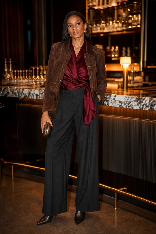 Burgundy Satin and Suede Evening Look A woman wears a brown suede jacket over a burgundy satin wrap top with black wide-leg trousers, pointed black boots, hoop earrings, and a black clutch.