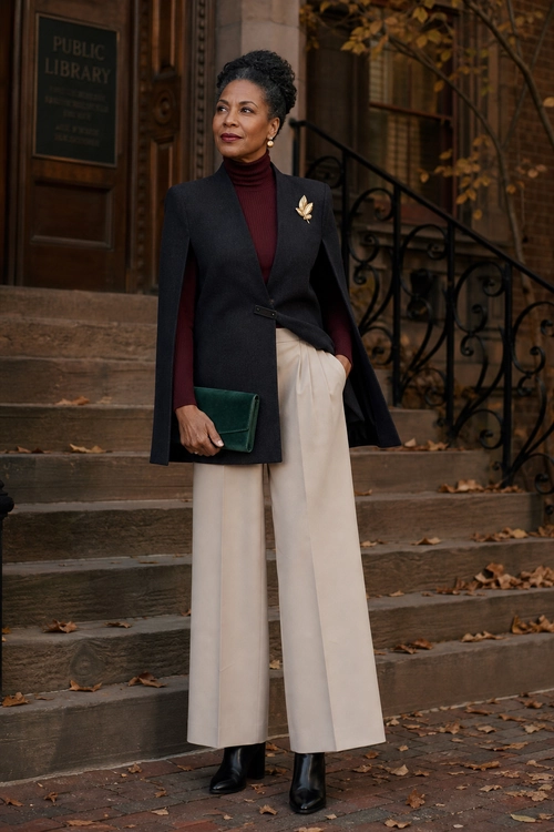 A woman wears a black cape-style blazer over a burgundy turtleneck with cream wide-leg trousers, black ankle boots, a green clutch, and a gold brooch.