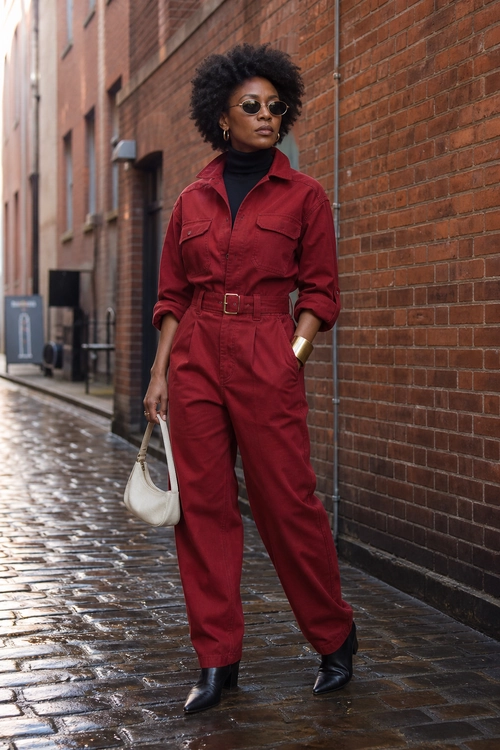 Burgundy Utility Jumpsuit Outfit A person wears a burgundy belted utility jumpsuit over a black turtleneck with black ankle boots, round sunglasses, and a white shoulder bag.