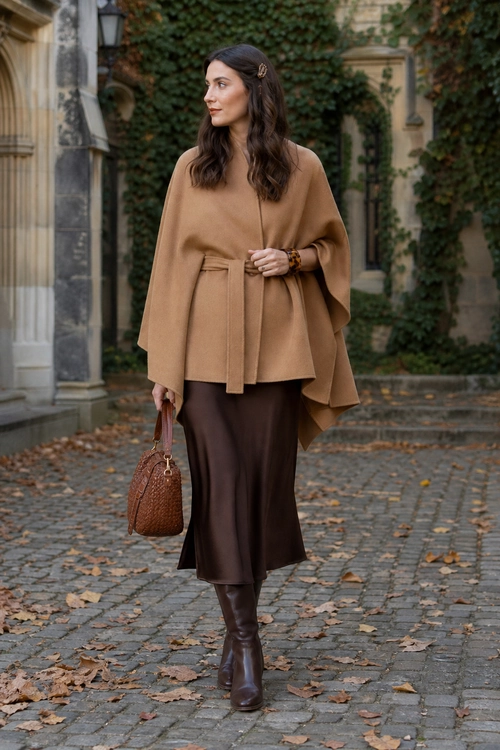 Camel Belted Poncho Outfit A woman wears a camel belted poncho with a dark brown midi skirt, tall boots, and a textured brown handbag.