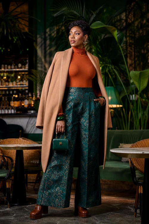 Camel Coat and Brocade Trousers A woman wears a camel coat draped over a rust turtleneck top with teal brocade wide-leg trousers, brown platform shoes, and a small green handbag.