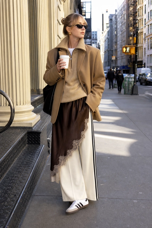 Camel Jacket and Layered Skirt Outfit A street-style outfit featuring a camel jacket, beige knit sweater, brown lace-trimmed skirt layered over cream trousers, white striped sneakers, sunglasses, and a black shoulder bag.