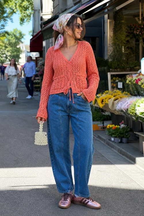 Coral Cardigan and Wide-Leg Jeans Outfit A street-style outfit featuring a coral open-knit cardigan, blue wide-leg jeans, a floral headscarf, sunglasses, a beaded handbag, and pink sneakers.
