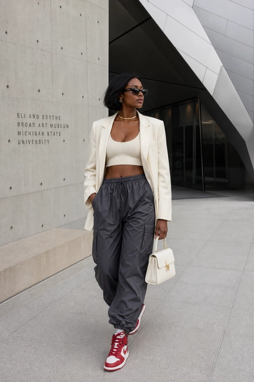 Cream Blazer and Gray Streetwear Outfit A fashion look featuring a cream blazer over a cropped cream top, charcoal gray pants, red and white sneakers, and a white handbag.