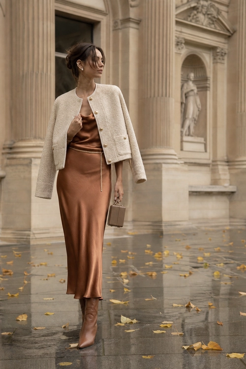 Cream Jacket and Copper Satin Dress A woman wears a cream cropped jacket over a copper satin midi dress with brown boots and a small structured handbag.