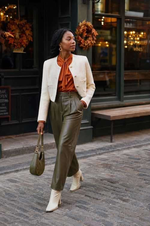 Cream Jacket and Olive Leather Trousers A woman wears a cream cropped jacket over a rust tie-neck blouse with olive leather trousers, cream ankle boots, gold earrings, and an olive handbag.