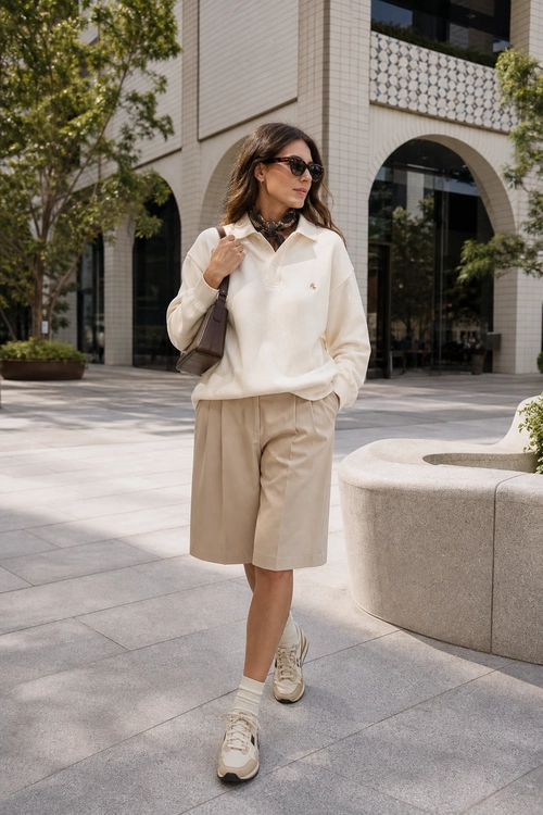 Cream Shirt and Tailored Bermuda Shorts A person wears a cream collared top with beige Bermuda shorts, white socks, light sneakers, sunglasses, and a shoulder bag outdoors.