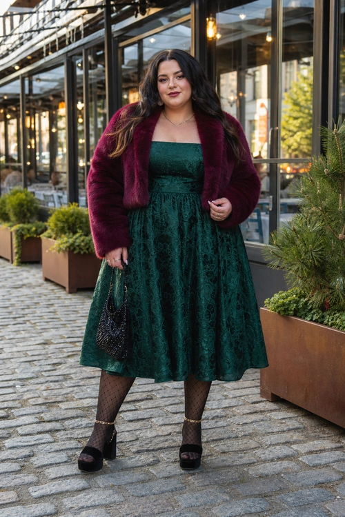 Emerald Dress with Burgundy Faux Fur A woman wears an emerald green midi dress with a burgundy faux fur jacket, black patterned tights, black platform heels, and a small black handbag.
