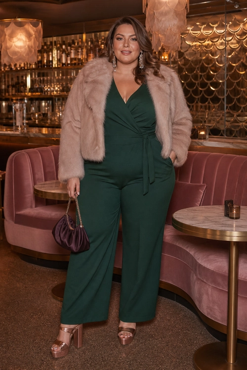 A woman wears an emerald green wrap-style wide-leg jumpsuit with a blush faux fur jacket, metallic platform sandals, drop earrings, and a purple handbag.
