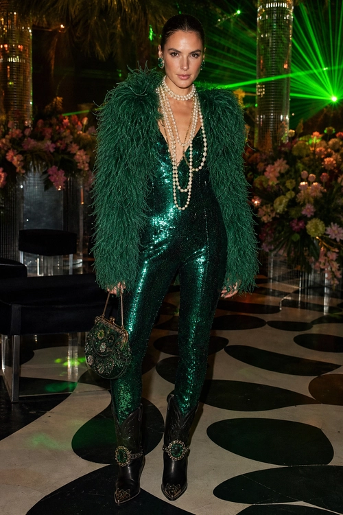 Emerald Sequin Jumpsuit Look A woman wears a dark green sequin jumpsuit with a feathered jacket, layered pearl necklaces, a decorative handbag, and embellished black boots.