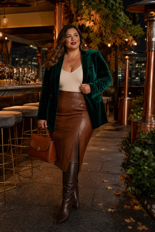A woman wears an emerald velvet blazer with a cream ribbed top, brown leather skirt, knee-high boots, and a structured handbag.