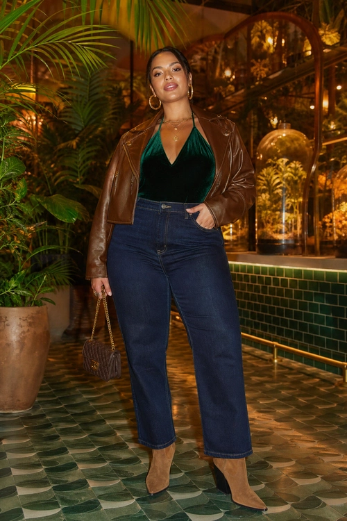 A woman wears an emerald green velvet halter top with a brown leather jacket, dark high-waisted jeans, tan ankle boots, and a small chain-strap handbag.