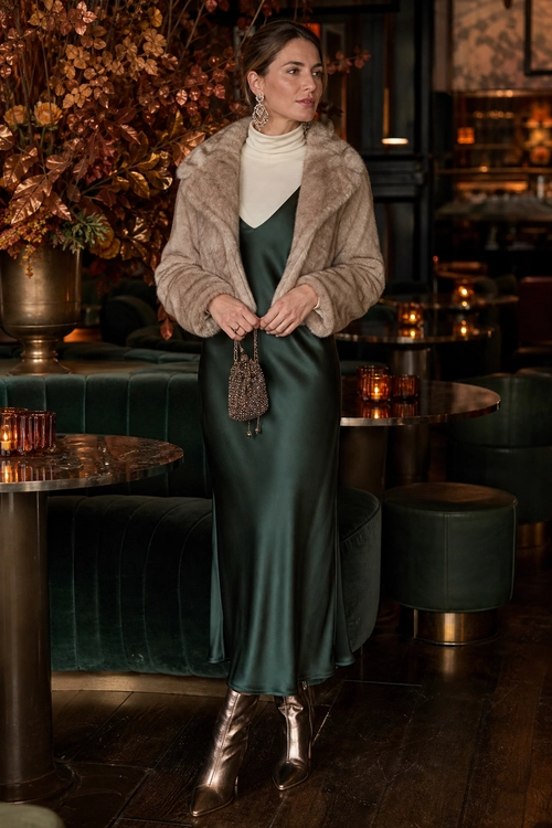 A woman wears a dark green satin slip dress layered over a cream turtleneck with a beige faux fur jacket, metallic ankle boots, statement earrings, and a small beaded bag.