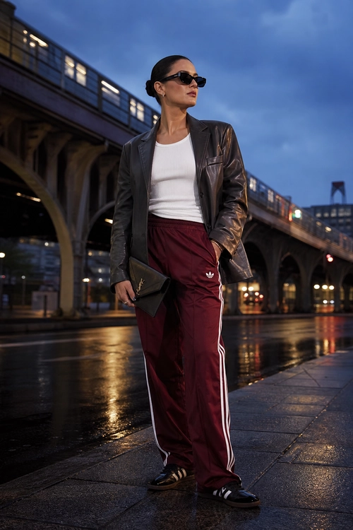 Leather Blazer and Burgundy Track Pants A woman wears a dark leather blazer over a white ribbed top with burgundy striped track pants, black sneakers, sunglasses, and a clutch.