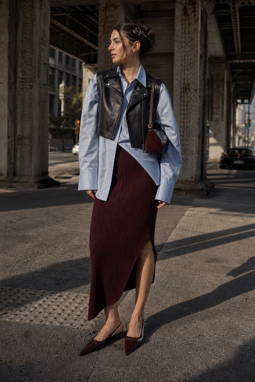 Leather Vest and Burgundy Skirt Look A woman wears a cropped black leather vest over a light blue shirt with a burgundy ribbed asymmetrical skirt, pointed shoes, and a small shoulder bag.