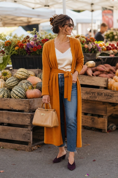 Mustard Cardigan Market Outfit A woman wears a long mustard tie-waist cardigan over a white V-neck top with blue jeans, dark pointed flats, sunglasses, and a tan top-handle handbag.